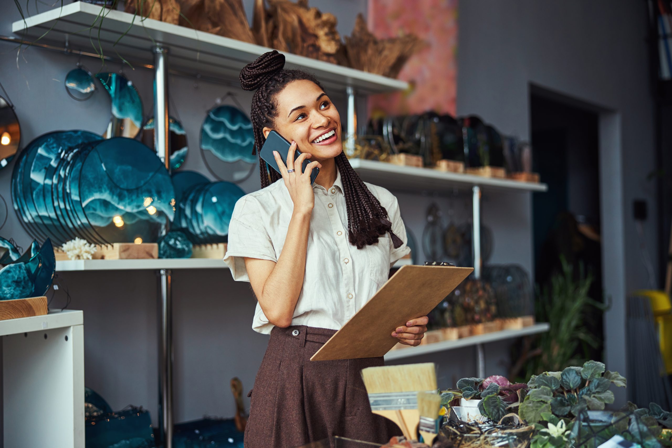 Smiling merry young floral designer with a clipboard in one hand talking on her smartphone